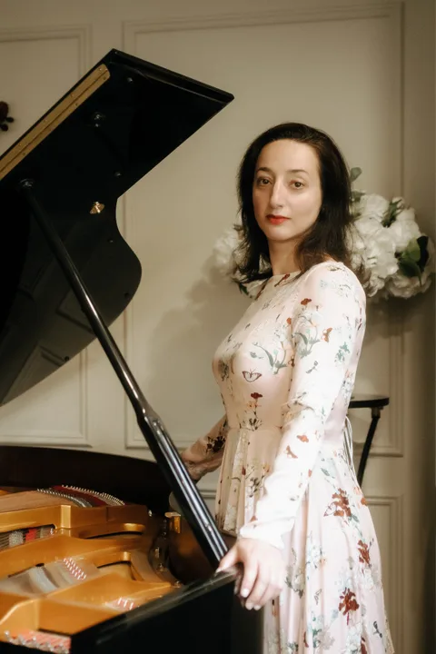 Mekhriban Mamedova standing beside an open grand piano in a floral gown, one hand resting on the raised lid, the gilded soundboard behind her.