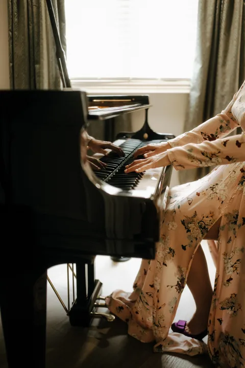Mekhriban Mamedova's hands on the keys of a black grand piano, seated in a floral gown by a curtained window.