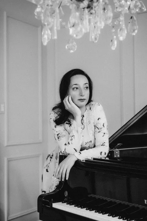 Black-and-white portrait of Mekhriban Mamedova leaning on the closed lid of a grand piano, chin resting on her hand, looking past the camera, crystal chandelier drops above her and paneled walls behind.