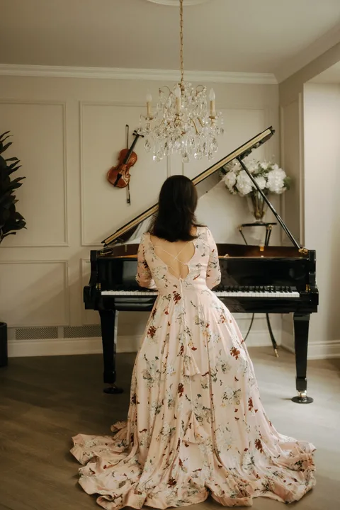 Mekhriban Mamedova seen from behind at a black grand piano, her floral gown trailing to the floor beneath a crystal chandelier.