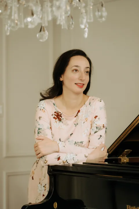 Mekhriban Mamedova leaning on the closed lid of a grand piano with a warm expression, looking to the side.