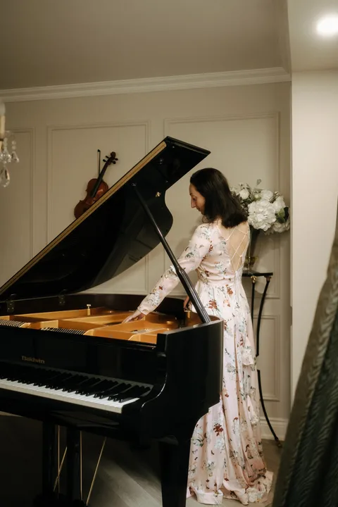 Mekhriban Mamedova standing at an open grand piano in a long floral gown, reaching in toward the strings, a violin hanging on the paneled wall behind her.