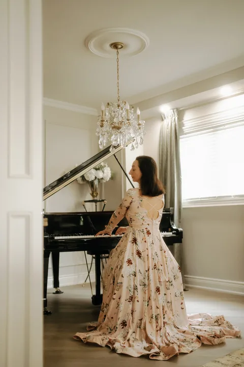 Mekhriban Mamedova in profile at the keyboard of a grand piano, framed by a doorway in a chandelier-lit room.
