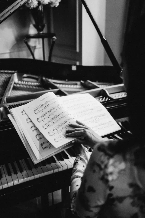 Black-and-white view over Mekhriban Mamedova's shoulder as she turns the pages of a bound score resting on the piano keys.