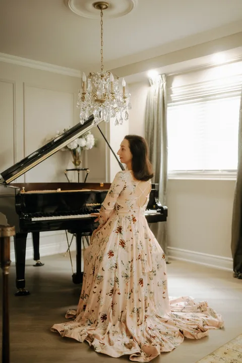 Mekhriban Mamedova seated at a black grand piano, turning toward the window light beneath a crystal chandelier.