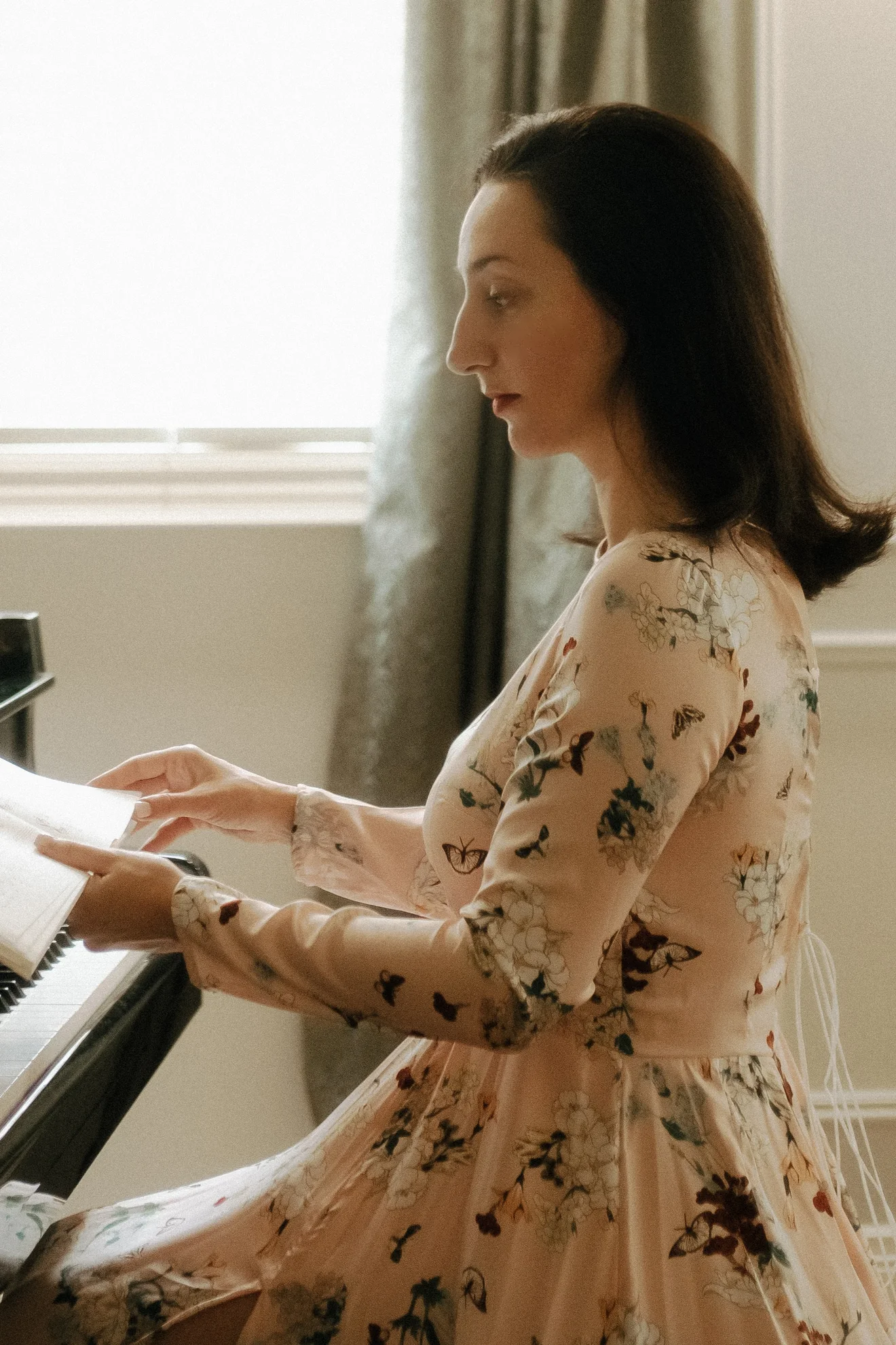 Mekhriban Mamedova seated in profile at an open grand piano, turning the pages of a score in the light of a tall window